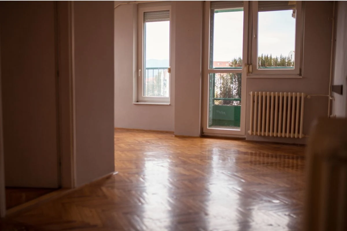 Puste mieszkanie z drewnianą podłogą, dużymi oknami balkonowymi oraz kaloryferem pod jednym z okien, do wnętrza wpada naturalne światło.