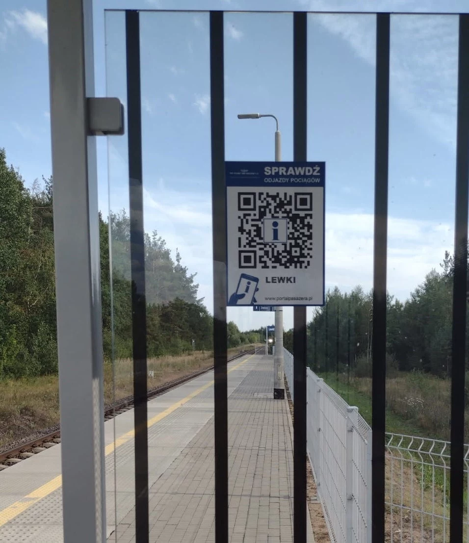 Przykładowy kod QR, które PKP PLK rozmieszcza na stacjach kolejowych. Tablica z kodem QR i napisem 'Sprawdź odjazdy pociągów' przyczepiona do szyby przystanku, w tle peron kolejowy, ogrodzenie oraz kawałek torów otoczonych drzewami.