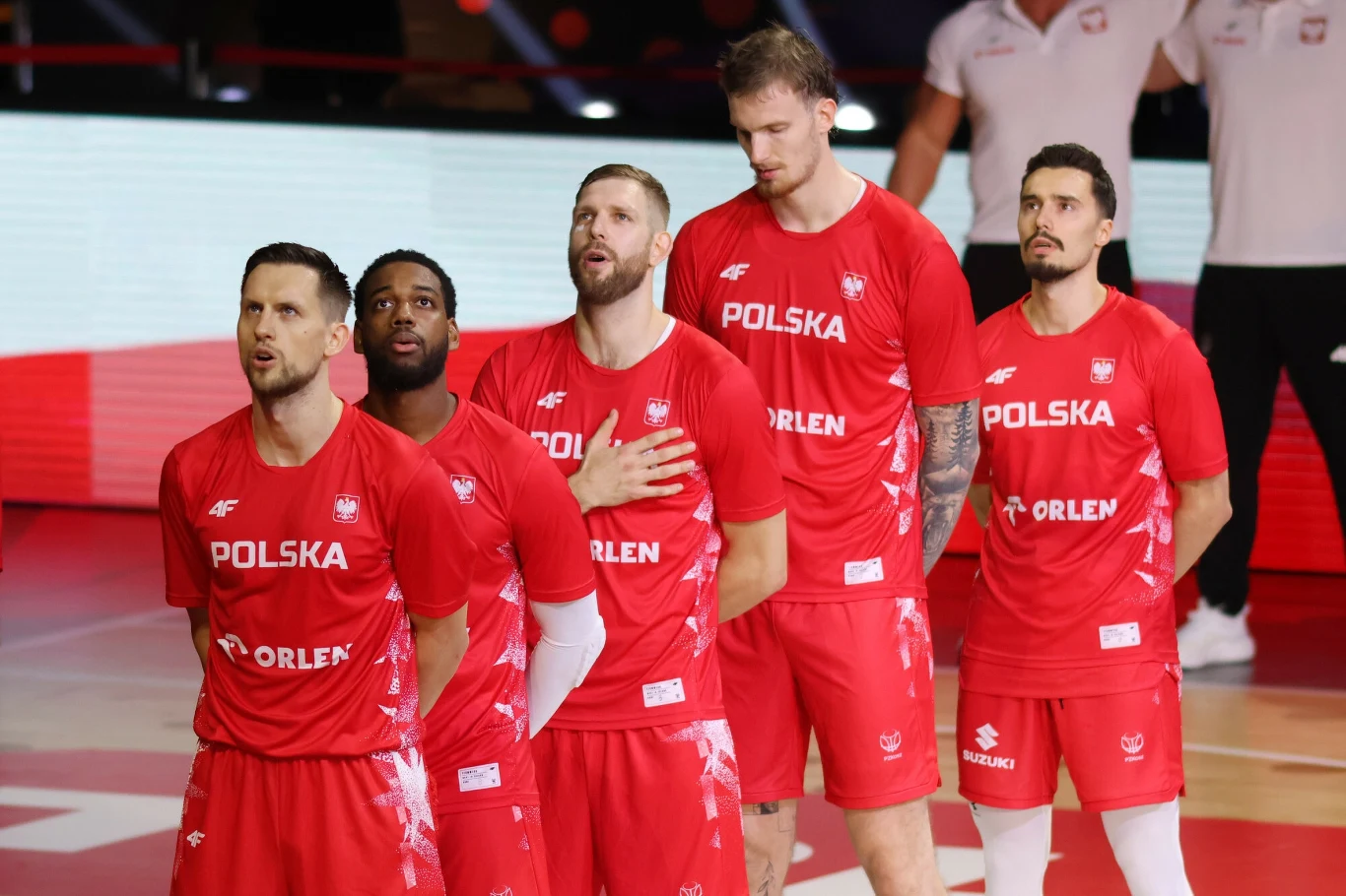 Grupa polskich siatkarzy ubranych w czerwone stroje reprezentacyjne stoi razem podczas oficjalnej ceremonii przed meczem, jeden z zawodników ma rękę położoną na sercu.