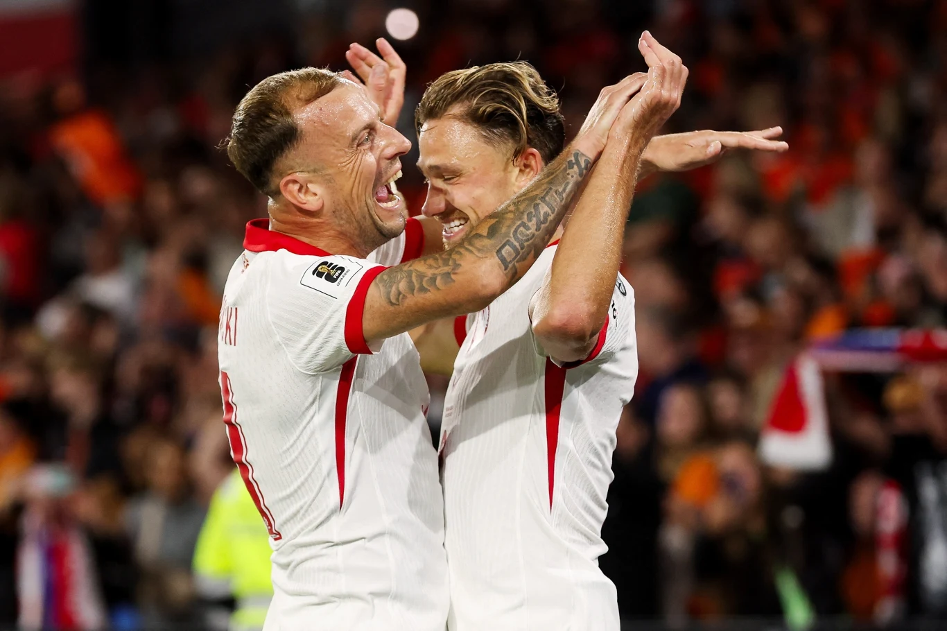 Kamil Grosicki, Matty Cash Dwóch piłkarzy w białych strojach cieszy się ze zdobytego gola na tle stadionu, jeden obejmuje drugiego, a wokół widać rozemocjonowanych kibiców.