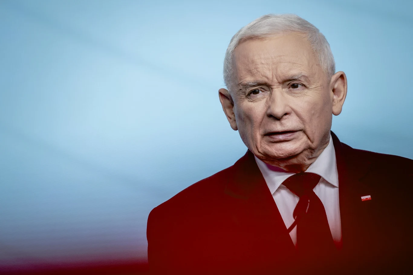 Czy prawica ma szansę na samodzielną władzę? Starszy mężczyzna o siwych włosach w garniturze i czerwonym krawacie, z niewielką flagą w klapie, na tle w odcieniach niebieskiego.