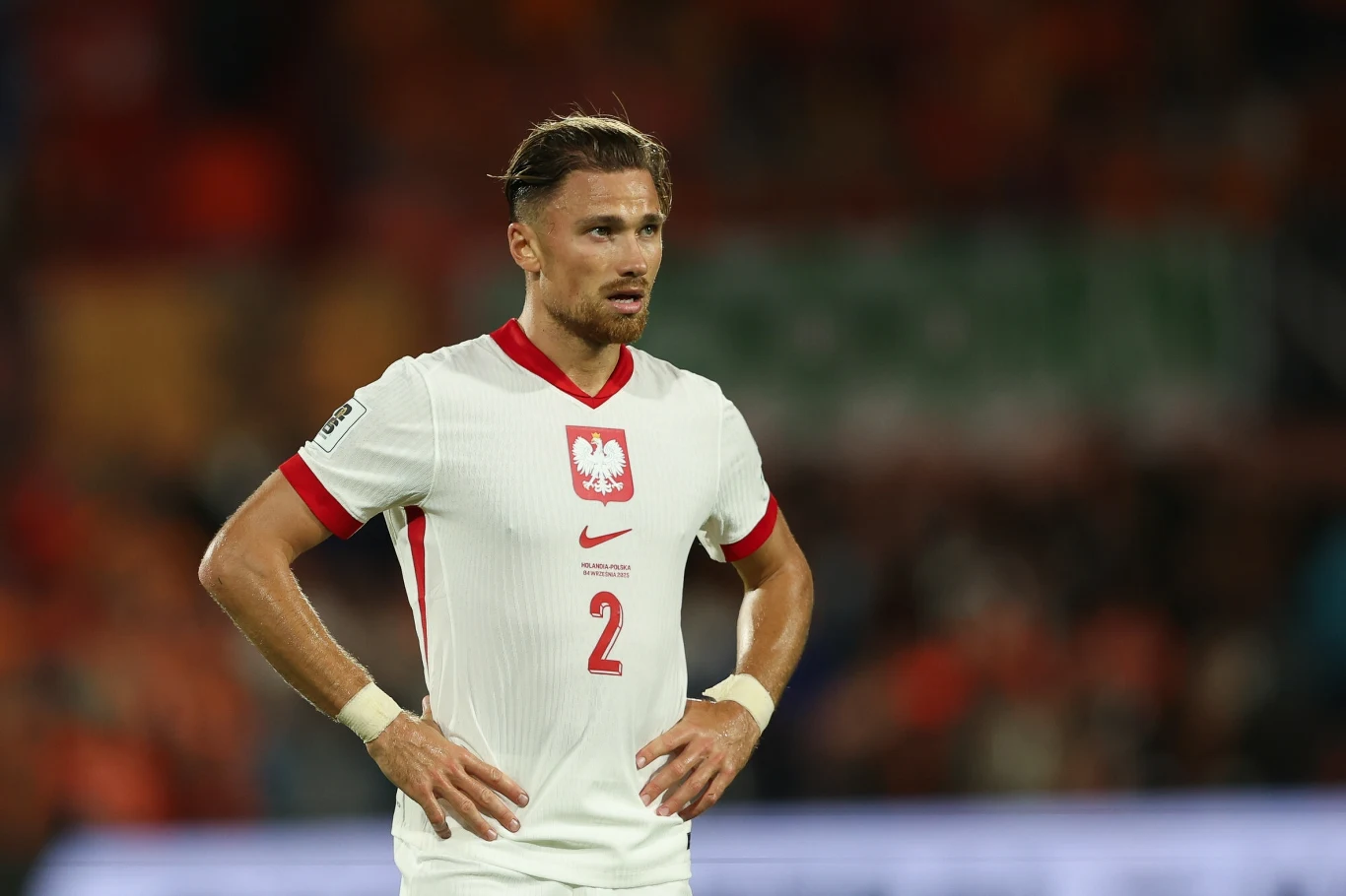 Matty Cash w meczu z Holandią Piłkarz w stroju reprezentacji Polski z numerem 2 na koszulce stoi na boisku podczas meczu, widoczny jest w skupionej pozie, z dłońmi opartymi na biodrach.