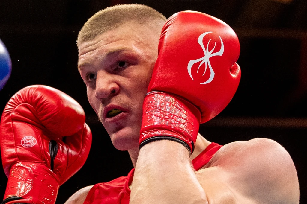 Adam Tutak Bokser w czerwonych rękawicach ochronnych podczas walki, z widocznym skupieniem na twarzy, podniesionymi rękami w obronnej pozycji, w tle ciemna sala sportowa.