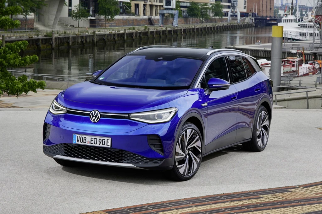 Volkswagen ID.4 jest dziś tańszy od Tiguana w podstawowej specyfikacji. Niebieski elektryczny SUV marki Volkswagen zaparkowany nad wodą, na tle nowoczesnej zabudowy miejskiej i jachtów. Lśniąca karoseria, charakterystyczne reflektory LED i efektowne aluminiowe felgi przyciągają uwagę, podkreślając dynamiczny charakter poja...