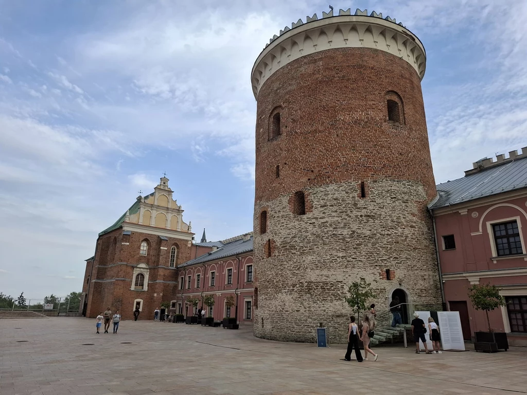 Duża, okrągła baszta z czerwonej cegły oraz przylegające do niej zabytkowe budynki o jasnych elewacjach, tło stanowi jasne niebo z delikatnym zachmurzeniem, w pobliżu spacerują ludzie.