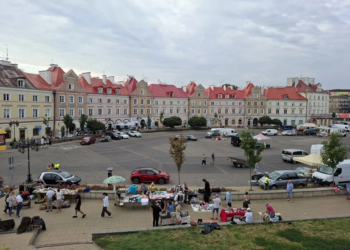 Rynek otoczony zabytkowymi kamienicami z charakterystycznymi czerwonymi dachami, na pierwszym planie stragany miejskiego targu oraz ludzie spacerujący i robiący zakupy