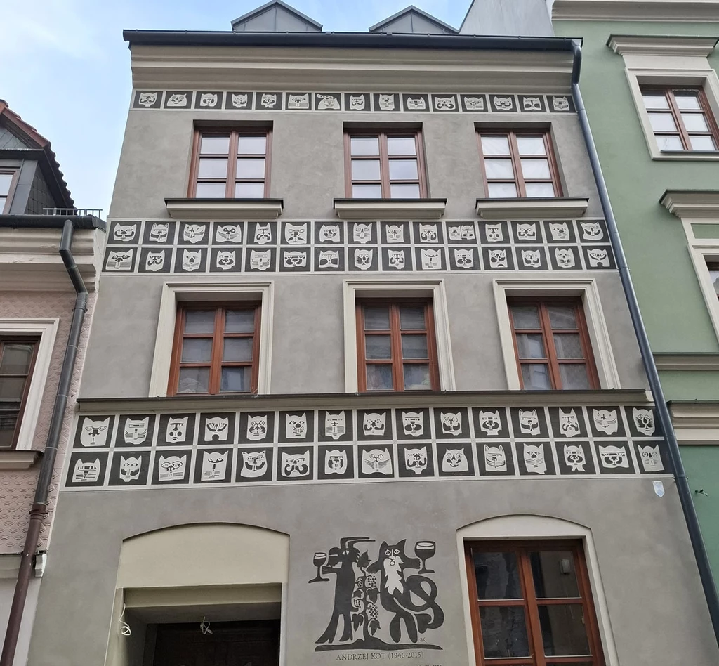 Kamienica z szarą elewacją ozdobiona prostokątnymi panelami z rysunkami kotów, na parterze umieszczona grafika z sylwetkami dwóch postaci trzymających kieliszki i napis 'Składzik Kresowy'.