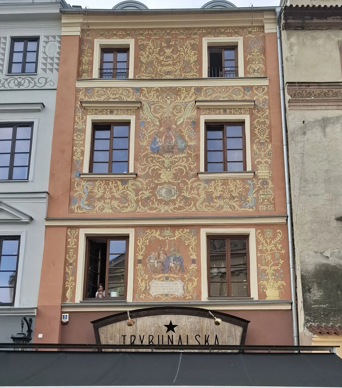 Fasada zabytkowej kamienicy z bogato zdobioną, kolorową polichromią i historycznymi motywami, cztery prostokątne okna, na parterze szyld Trybunalska, ozdoby roślinne i figuralne akcenty podkreślające historyczny charakter budynku.