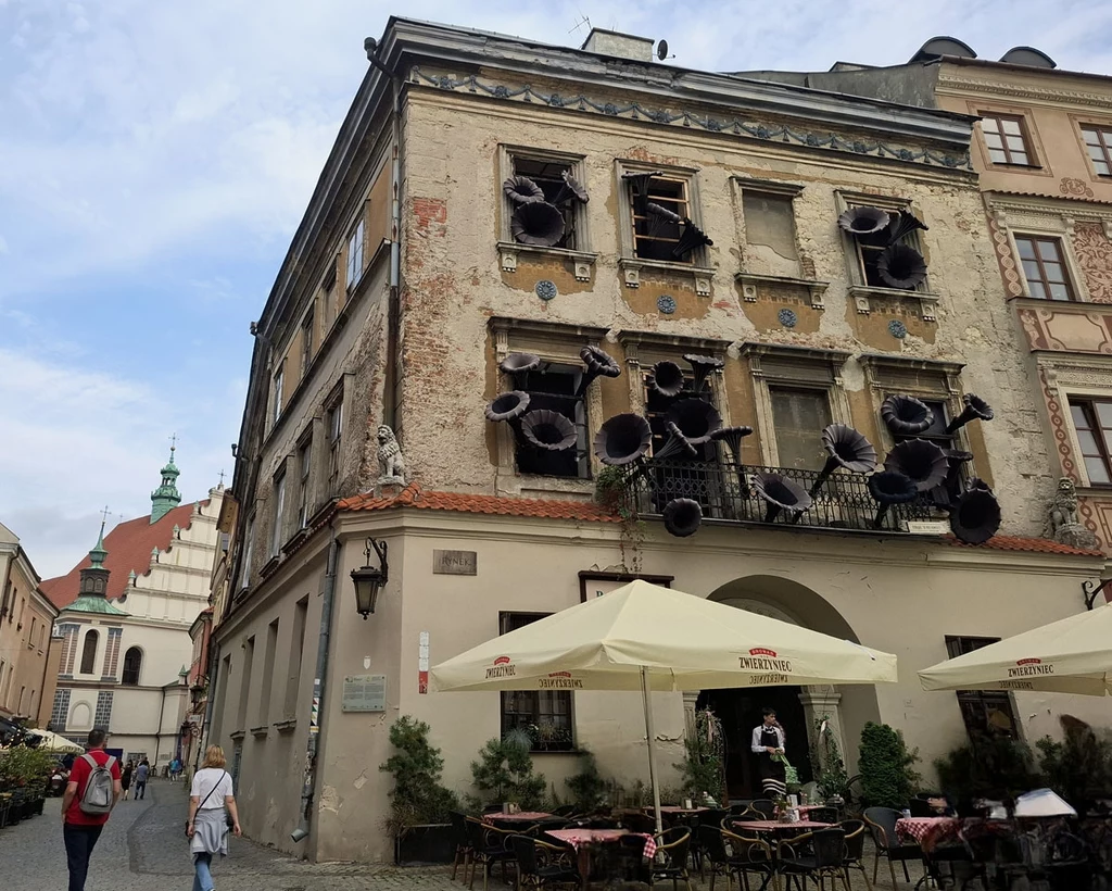 Stara kamienica z odpadającym tynkiem, na której oknach i elewacji zamontowano liczne duże czarne megafony, przed budynkiem stoi ogródek kawiarniany z jasnymi parasolami, a ulicą przechodzą ludzie.