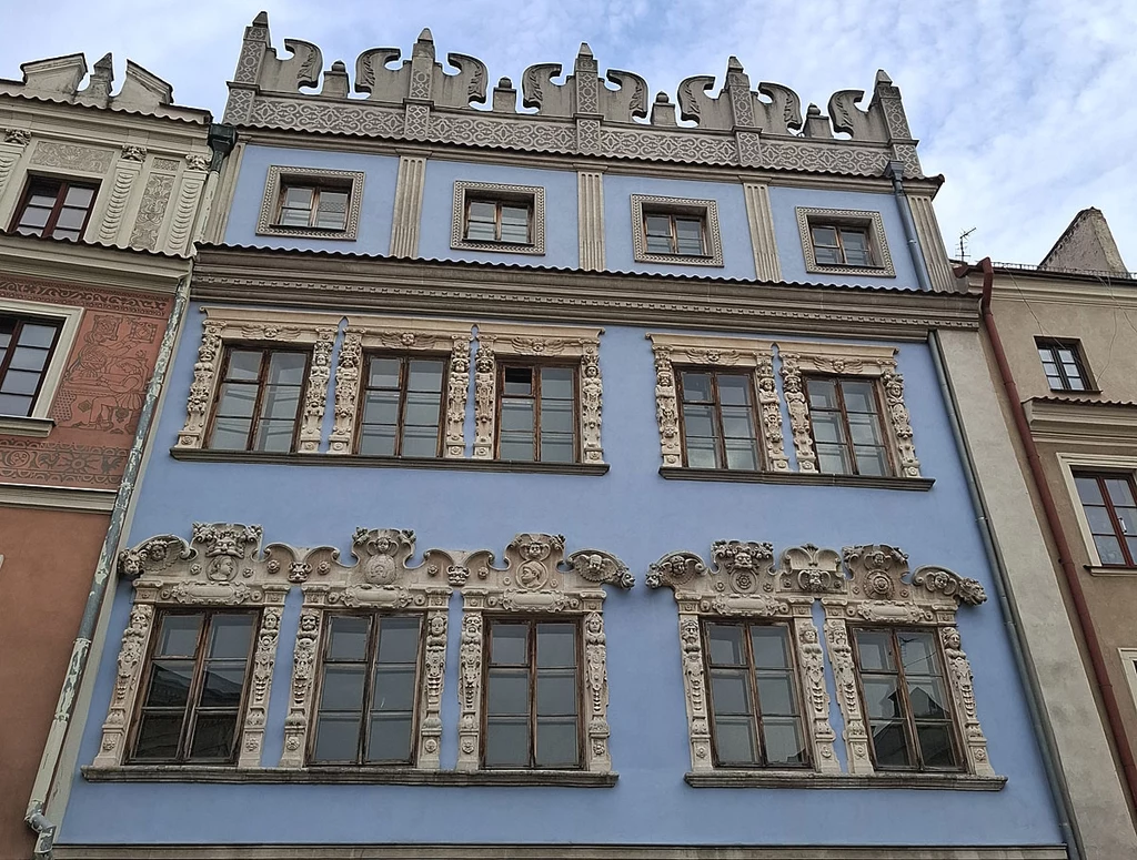Fasada historycznej kamienicy o błękitnym kolorze z dekoracyjnymi sztukateriami wokół okien, bogato zdobiony szczyt i elementy renesansowe, sąsiadujące budynki widoczne po bokach.