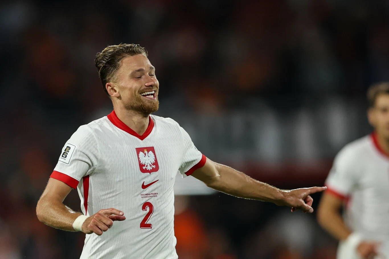 Matty Cash w meczu Holandia - Polska Polski piłkarz w białej koszulce z orłem i numerem 2, uśmiechnięty, biegnący po murawie podczas meczu piłkarskiego, drugi zawodnik widoczny jest w tle.