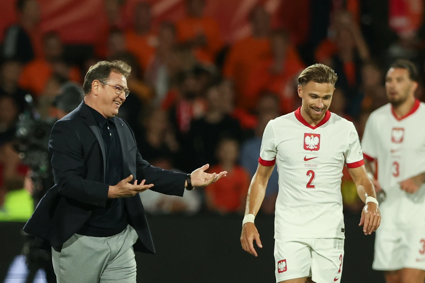Jan Urban i Matty Cash podczas meczu Holandia - Polska (4 września 2025) Trener w garniturze rozmawia z piłkarzem reprezentacji Polski ubranym w biały strój z czerwonymi akcentami, na tle kibiców w pomarańczowych barwach.