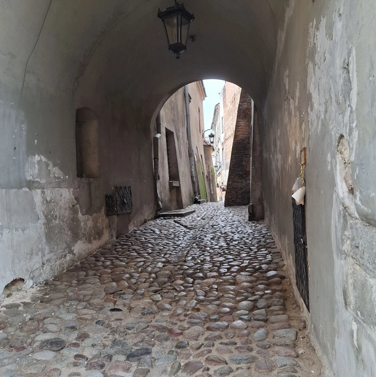 Kamienna brukowana uliczka przechodząca przez wąski, historyczny tunel z widoczną lampą wiszącą pod sklepieniem, otoczona starymi, otynkowanymi ścianami z wyraźnymi śladami upływu czasu.