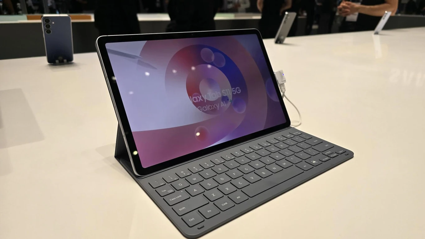 Tablet Samsung Galaxy z klawiaturą w etui, wyeksponowany na jasnym blacie, w tle rozmazane sylwetki kilku osób oraz inne urządzenia elektroniczne.