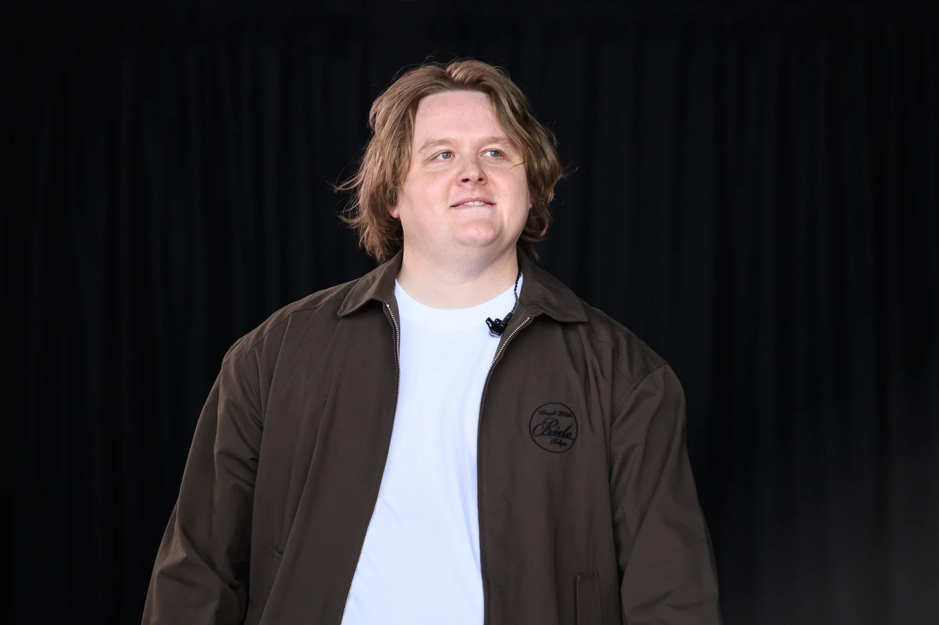 Lewis Capaldi podsumował dzisiejszą branżę muzyczną Młody mężczyzna o jasnych włosach w białej koszulce i brązowej kurtce stoi na tle ciemnej kotary, patrzy lekko w górę i uśmiecha się subtelnie.