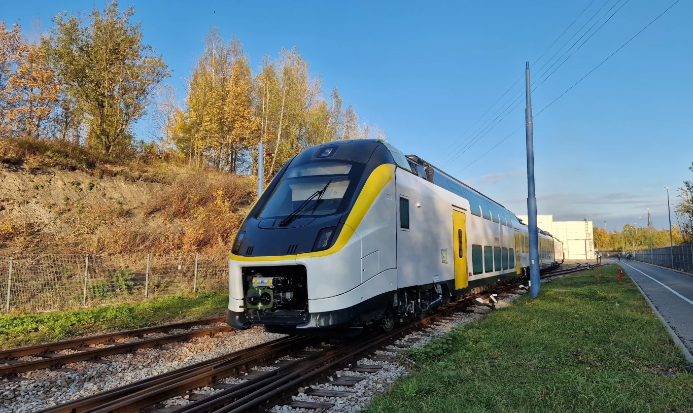 Pociągi piętrowe dla PKP Intercity. Alstom ujawnia nowe szczegóły. Dwupiętrowy nowoczesny pociąg o żółtych i białych barwach jadący po torach obok nasypu porośniętego jesiennymi drzewami, pogodny dzień z niebieskim niebem
