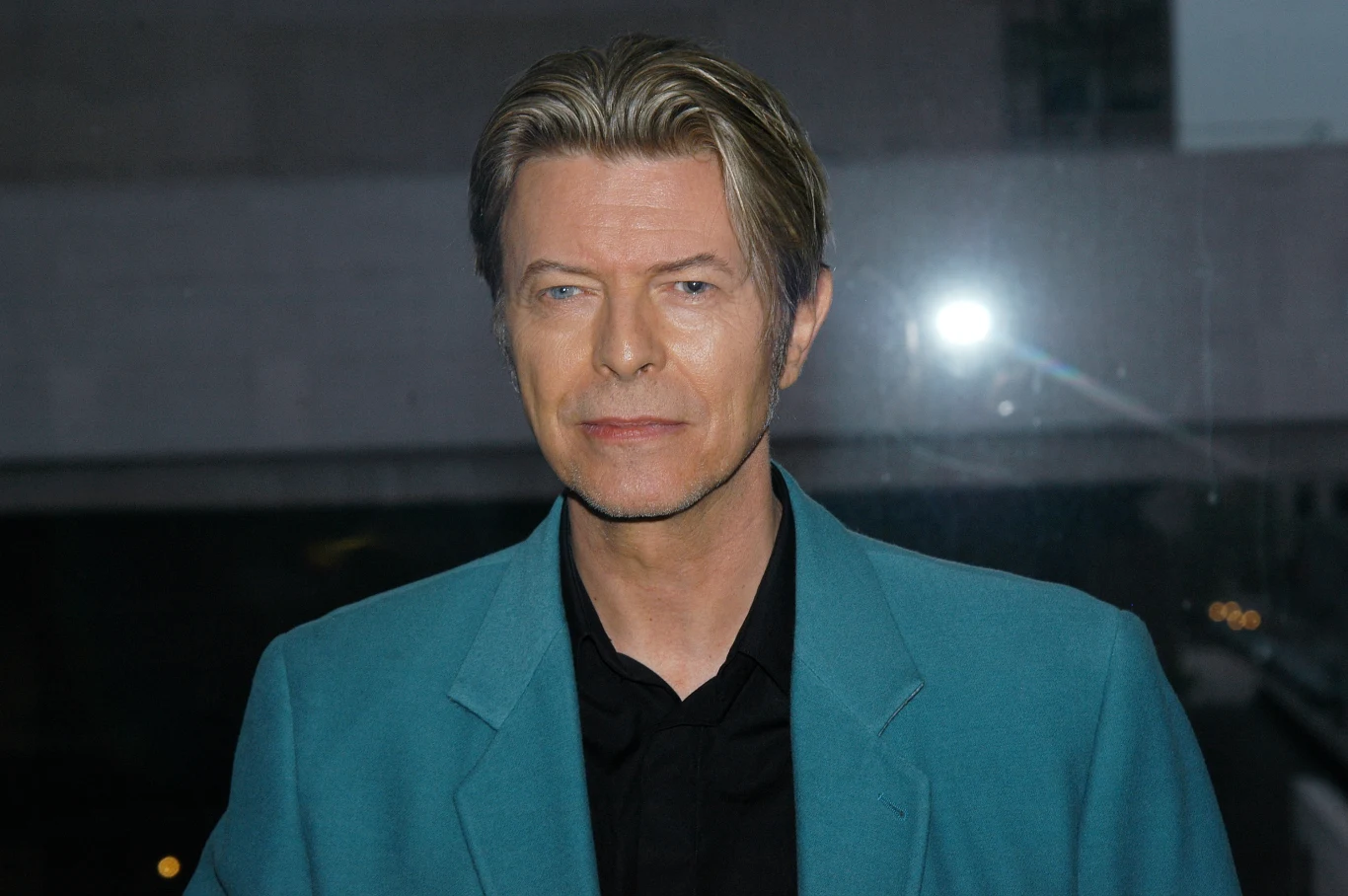 David Bowie Mężczyzna z charakterystycznymi, jasnymi włosami w niebieskiej marynarce oraz czarnej koszuli stojący na tle ciemnego, rozmytego otoczenia z widocznym błyskiem flesza.