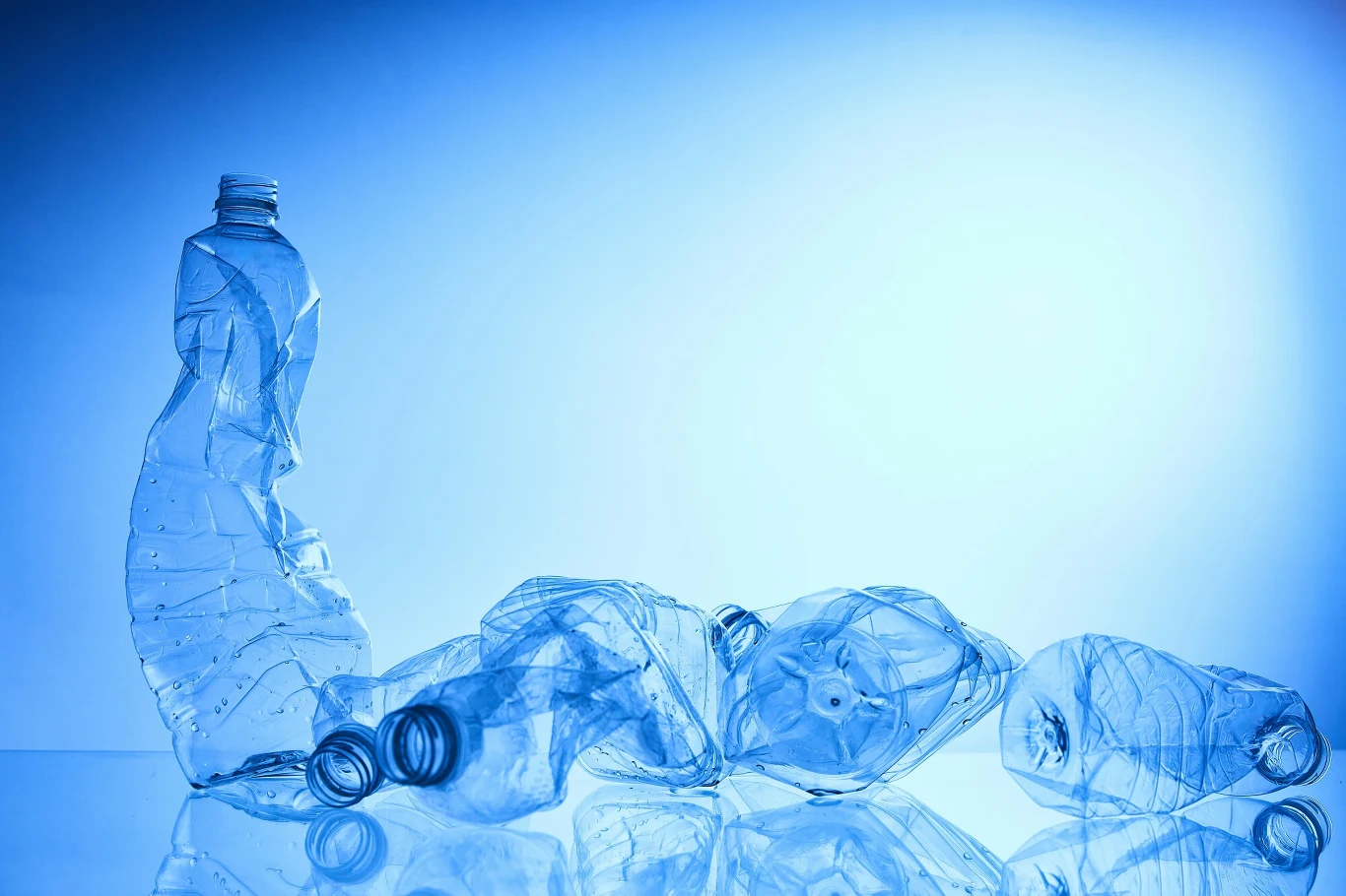 Kilka zgniecionych plastikowych butelek leżących na przezroczystej powierzchni w odcieniach niebieskiego światła. Najbardziej wyróżnia się jedna pionowa butelka, podczas gdy pozostałe leżą poziomo.