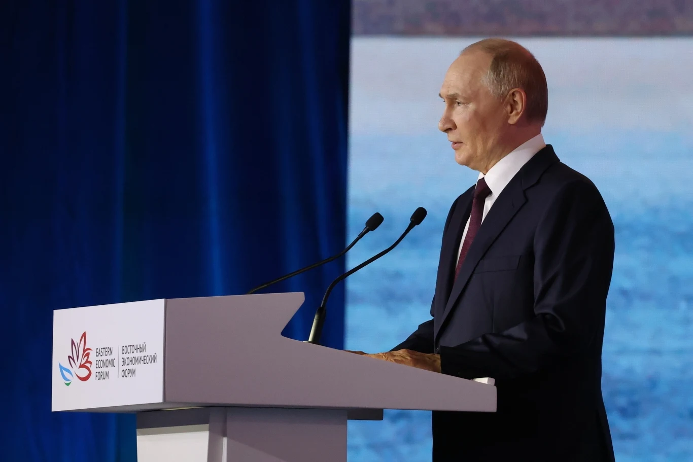 Władimir Putin podczas sesji plenarnej forum ekonomicznego we Władywostoku Mężczyzna w garniturze i krawacie przemawia przy mównicy, tło w odcieniach niebieskiego, widoczne mikrofony oraz logo i napisy na mównicy sugerujące konferencję lub forum ekonomiczne.