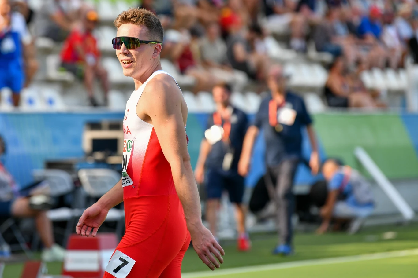 Jakub Szymański Sportowiec w czerwono-białym stroju startowym z numerem startowym 7 na torze lekkoatletycznym, uśmiechnięty, w okularach przeciwsłonecznych, na tle rozmytej publiczności oraz obsługi technicznej zawodów.