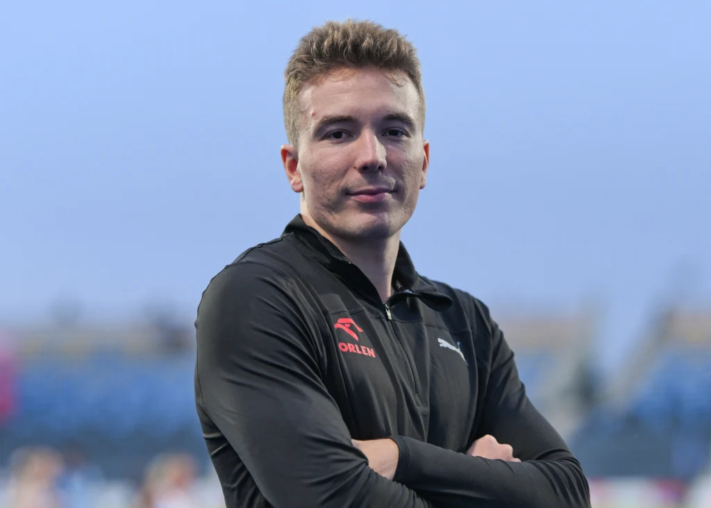 Jakub Szymański Młody mężczyzna w sportowym stroju z logotypem, ze skrzyżowanymi ramionami, stoi na tle nieostrego, zewnętrznego stadionu, patrząc bezpośrednio w obiektyw.