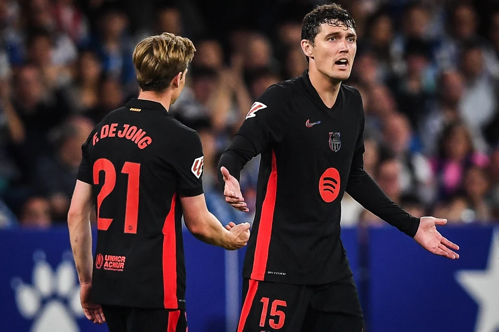 Frenkie de ong i Andreas Christensen Dwóch piłkarzy drużyny w czarnych strojach z czerwonymi akcentami podczas meczu, jeden z zawodników z numerem 15 gestykuluje w kierunku kolegi z zespołu, w tle rozmazane trybuny z kibicami.