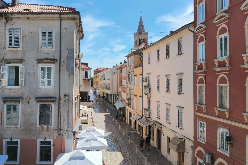 Dlaczego warto odwiedzić Zadar? Powodów jest wiele Malownicza, wąska uliczka wyłożona kostką brukową otoczona wysokimi, pastelowymi i historycznymi kamienicami, białe parasole ogródków kawiarnianych rozłożone wzdłuż ulicy, w tle widoczna wieża kościelna na tle błękitnego nieba.