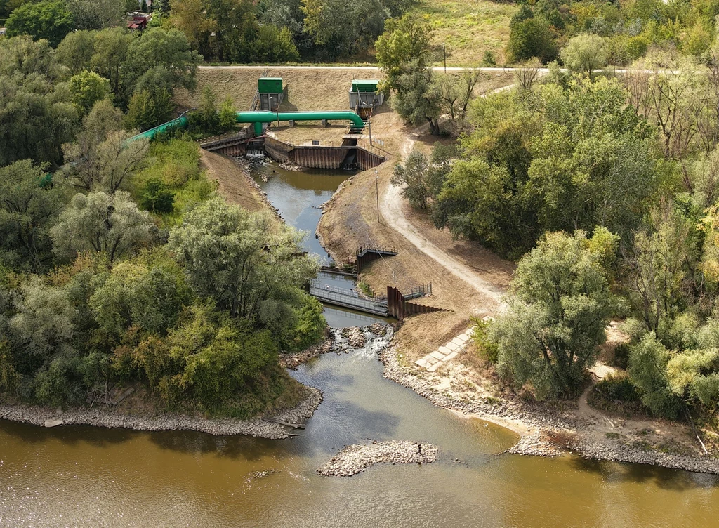 Kanał wyrzutowy ciepłej wody z elektrociepłowni Siekierki Rzeka otoczona drzewami z widoczną infrastrukturą hydrotechniczną, taką jak śluza i budowle wodne, pośród zielonego krajobrazu i nasypów ziemnych.