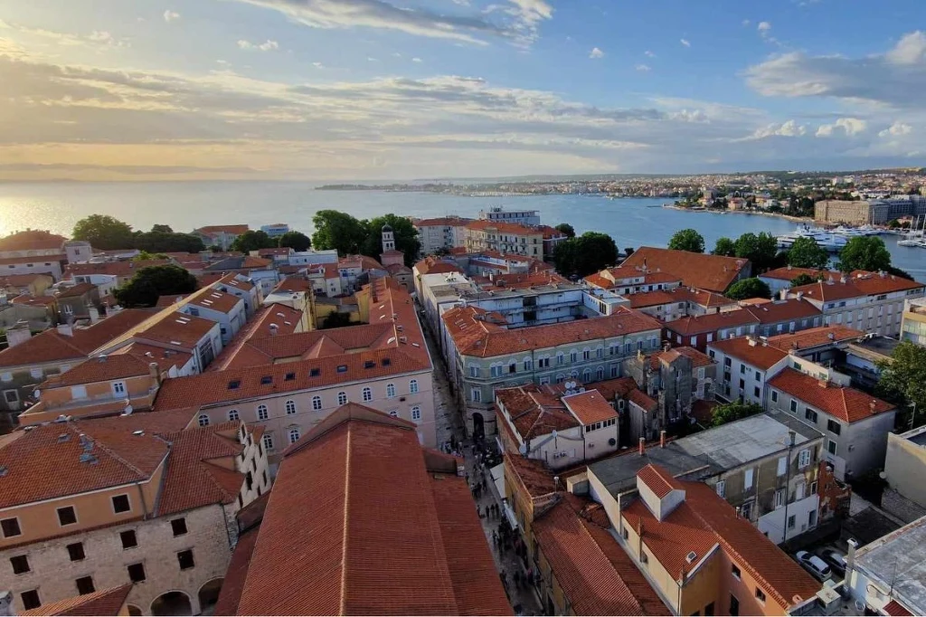 Zobacz widoki, które oczarowały światowej sławy reżysera Panorama miasta z licznymi czerwonymi dachami zabytkowych budynków, otoczonymi drzewami, z widokiem na morze i zachodzące słońce, w tle linia brzegowa z dalszą częścią miasta.