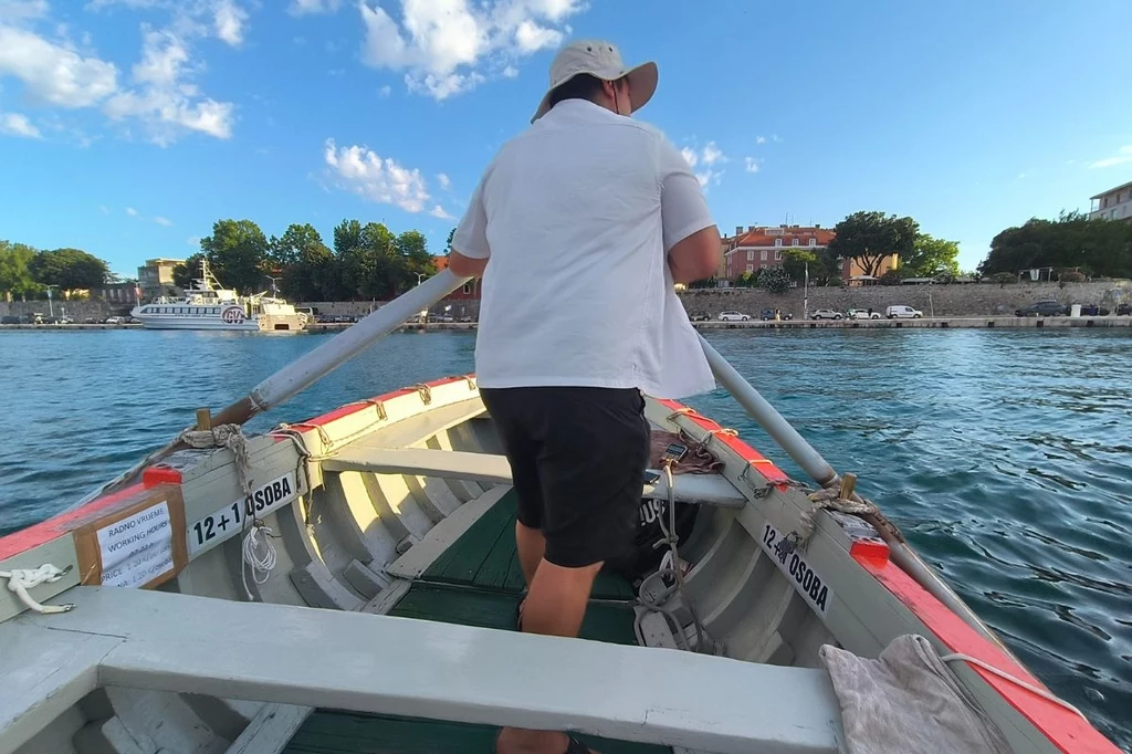 Komunikacja zbiorowa ma w Zadarze nieco inny wymiar Osoba w białej koszuli i kapeluszu wiosłująca w łodzi na tle spokojnej wody i zabudowy miejskiej w oddali, pod jasnym niebem z kilkoma chmurami.