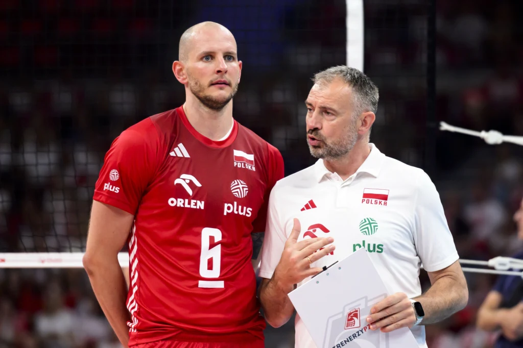 Bartosz Kurek i Nikola Grbić Siatkarz w czerwonym stroju numer 6 oraz trener w białej koszulce z logo Polskiego Związku Piłki Siatkowej omawiają taktykę podczas meczu siatkarskiego, w tle widoczne trybuny i siatka