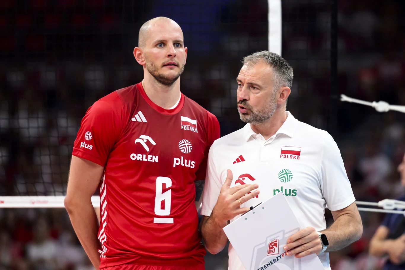 Siatkarz w czerwonym stroju numer 6 oraz trener w białej koszulce z logo Polskiego Związku Piłki Siatkowej omawiają taktykę podczas meczu siatkarskiego, w tle widoczne trybuny i siatka