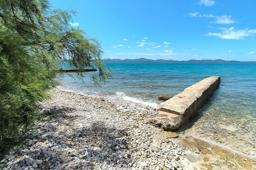 Plaże w Zadarze nie są szerokie, ale wygrywają spokojem Kamienista plaża z widokiem na lazurową wodę, na pierwszym planie betonowy pomost prowadzący do morza, po lewej stronie gałęzie zielonego drzewa, w tle pasmo gór oraz błękitne niebo z delikatnymi chmurami.