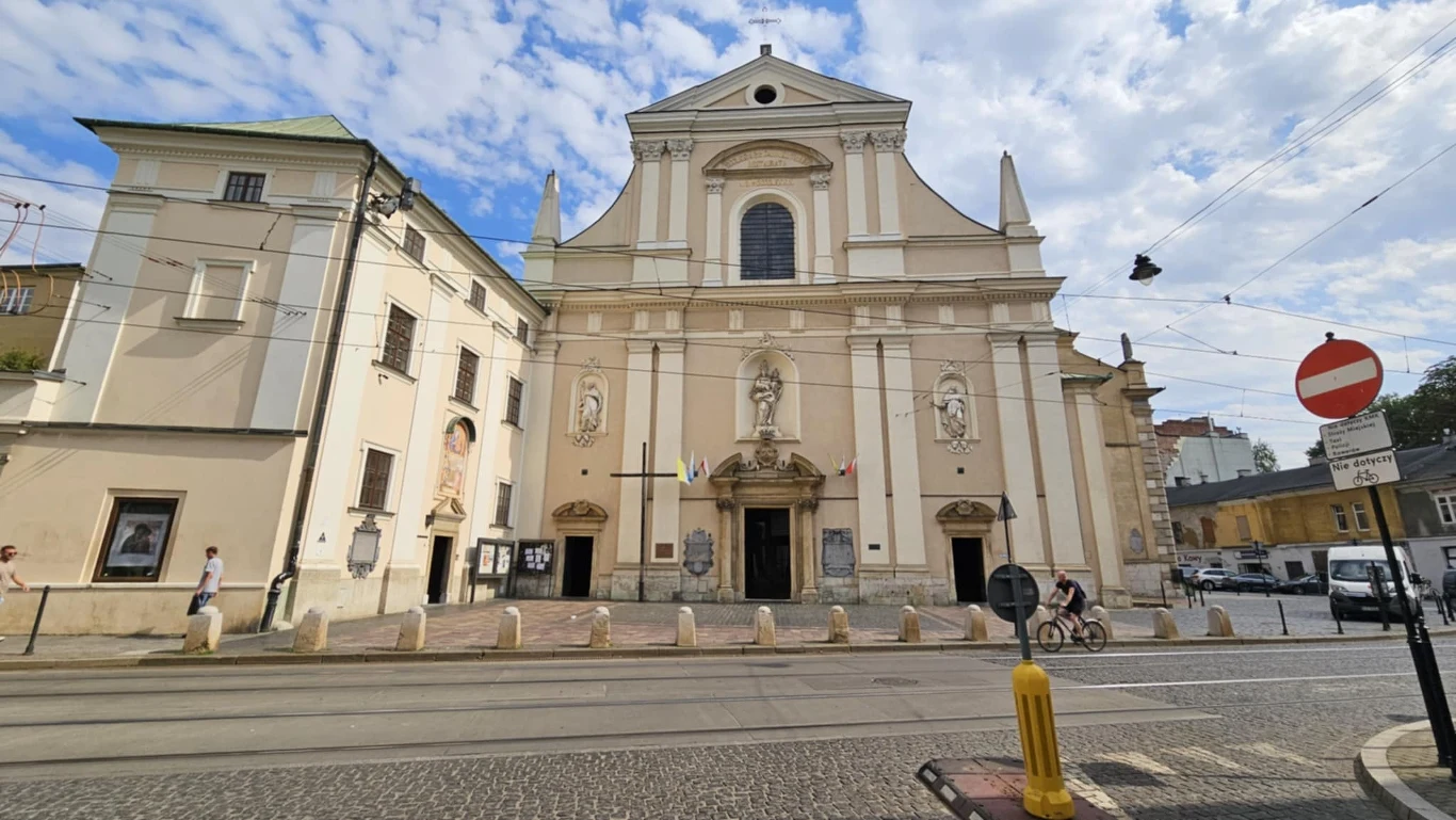 Duży, barokowy kościół z jasną fasadą ozdobioną trzema figurami świętych, dwoma bocznymi portalami oraz głównym wejściem, położony przy brukowanej ulicy z torowiskiem tramwajowym i znakami drogowymi, otoczony niskimi budynkami oraz z niewielką liczbą p...