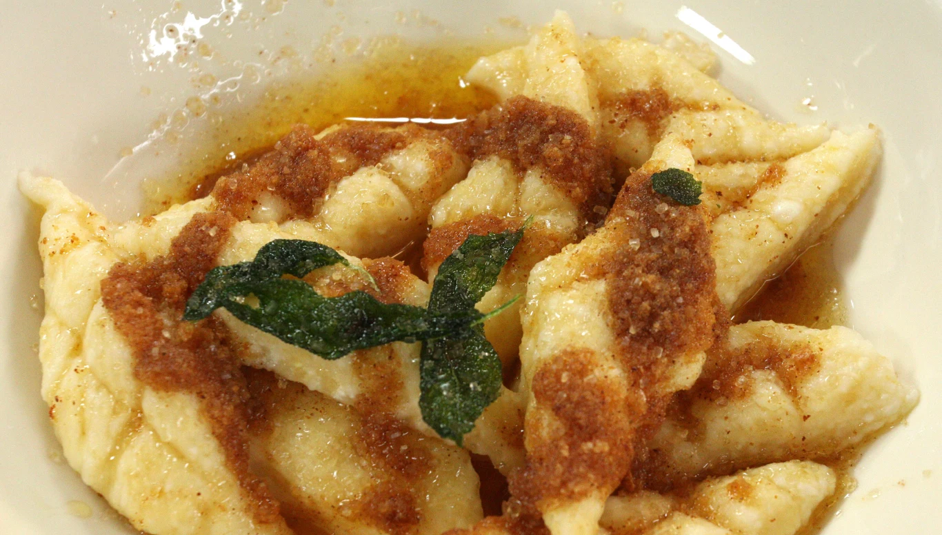 Leniwe z masłem szałwiowym Porcja kopytek poląwanych masłem zrumienionym z dodatkiem bułki tartej oraz świeżym liściem zioła, podane na białym talerzu.