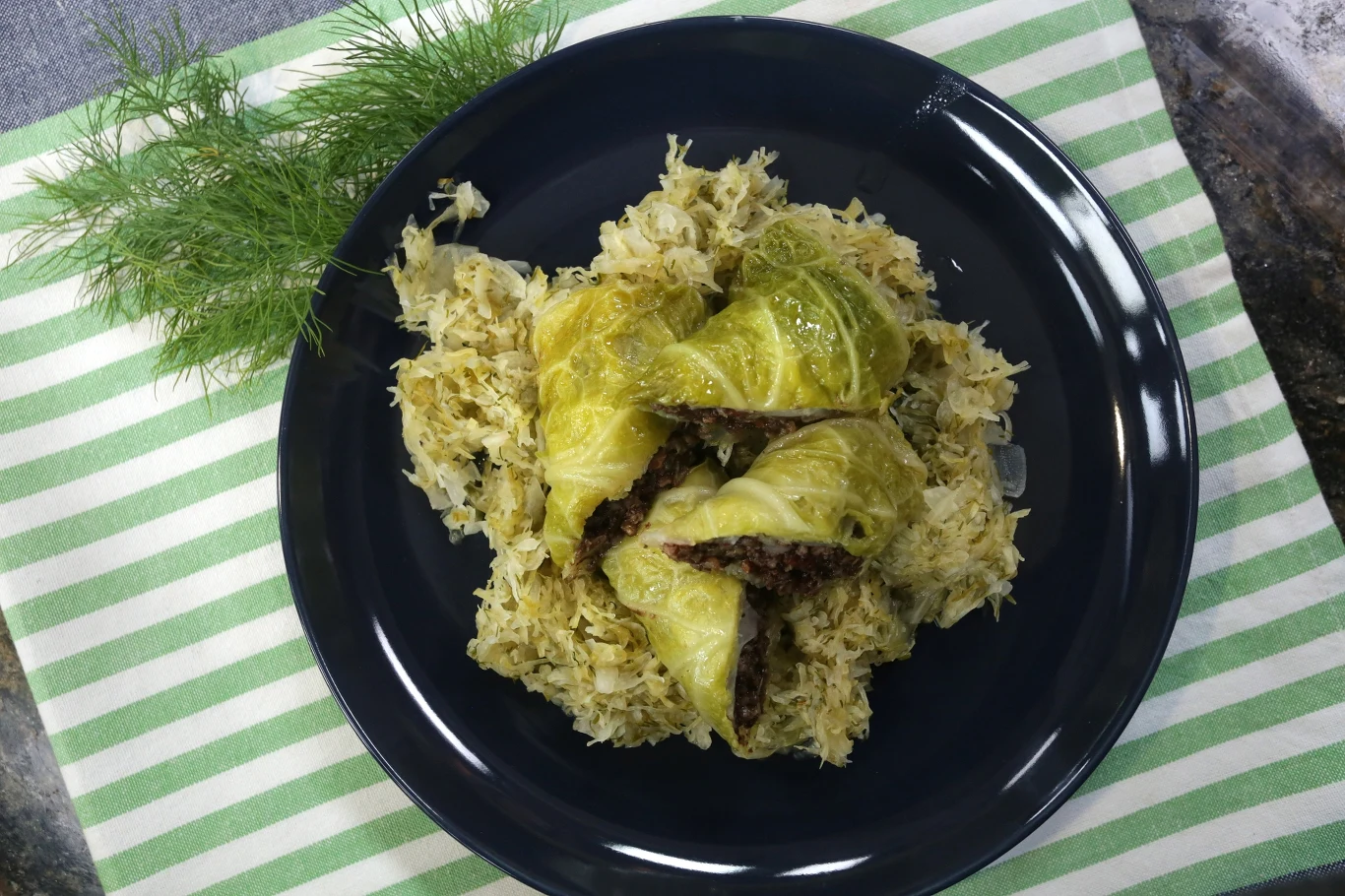 Gołąbki z kaszanką Gołąbki podane na duszonej kapuście na ciemnym talerzu, ułożonym na biało-zielonej ściereczce, obok świeży koper jako dekoracja.