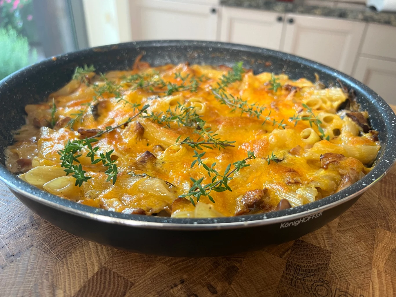 Zapiekanka makaronowa z kurkami Makaron zapiekany w naczyniu żaroodpornym, pokryty warstwą rozpuszczonego sera i udekorowany świeżym tymiankiem, otoczony jasnym, nowoczesnym wnętrzem kuchni.