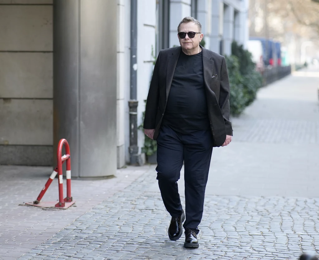 Mężczyzna w czarnych ubraniach i okularach przeciwsłonecznych spaceruje po brukowanym chodniku przy miejskiej ulicy, obok budynków i niewielkich roślin ozdobnych.