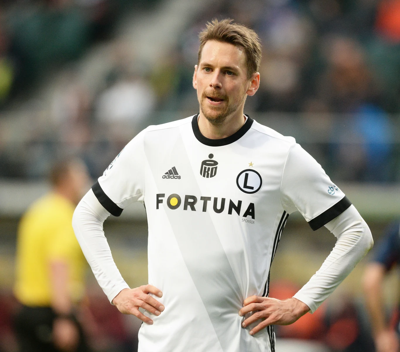 Piłkarz w białym stroju drużyny Legia Warszawa stoi na murawie stadionu z rękami opartymi na biodrach, w tle zamazane sylwetki innych osób i trybuny.
