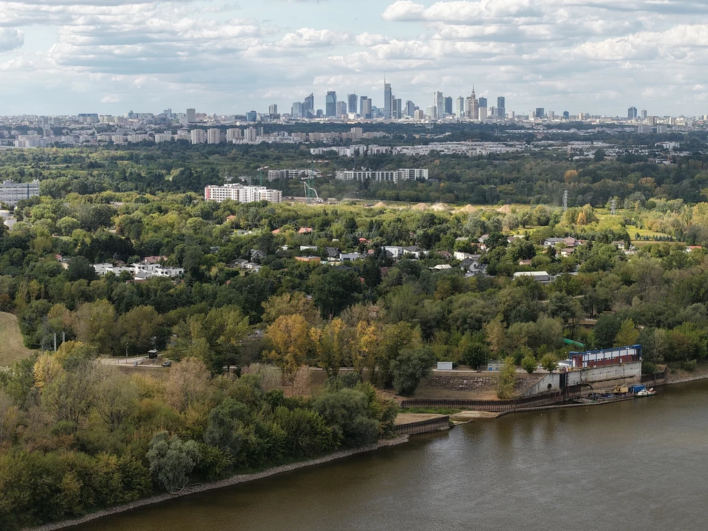 Przyczyną rekordowo niskich stanów wody w warszawskiej Wiśle jest nie tylko susza hydrologiczna Panorama miasta z widocznym centrum biurowym Warszawy w oddali, liczne zielone tereny i drzewa w centralnej części kadru, w pierwszym planie rzeka oraz zabudowania portowe.
