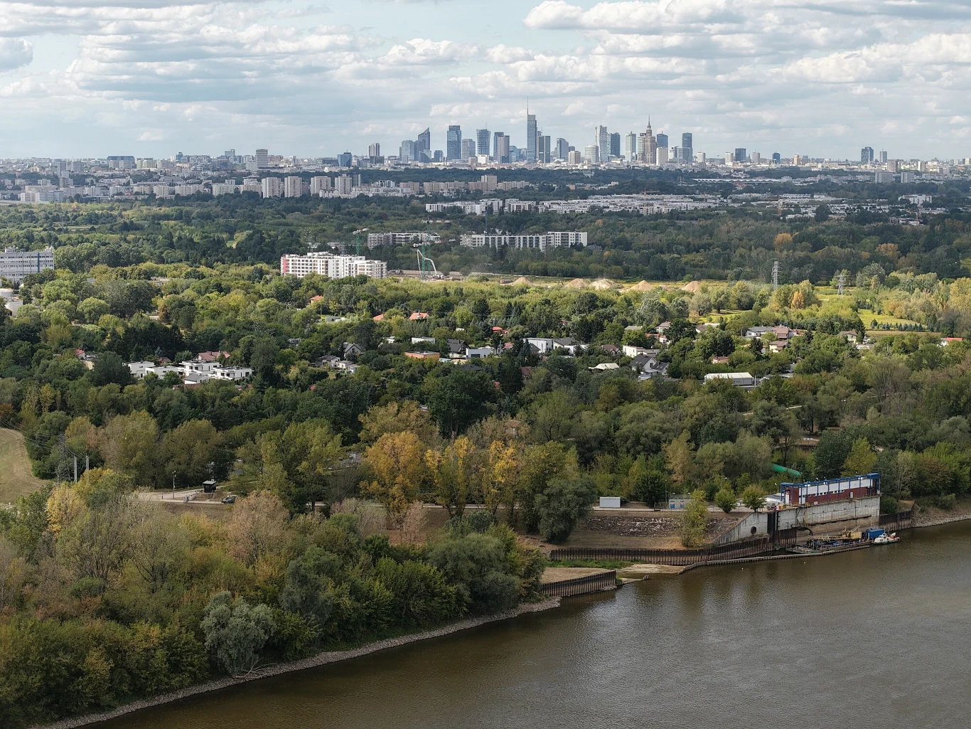 Przyczyną rekordowo niskich stanów wody w warszawskiej Wiśle jest nie tylko susza hydrologiczna Panorama miasta z widocznym centrum biurowym Warszawy w oddali, liczne zielone tereny i drzewa w centralnej części kadru, w pierwszym planie rzeka oraz zabudowania portowe.