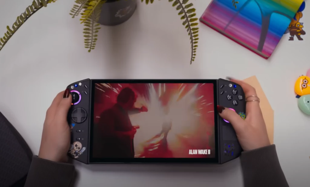 Konsola przenośna trzymana w dłoniach, na ekranie widoczna dynamiczna scena z gry Alan Wake II, w tle na biurku roślina doniczkowa, kolorowy notes oraz drobne akcesoria.