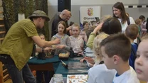 Grupa dzieci zgromadzona wokół stołu z materiałami edukacyjnymi, nauczyciel demonstruje przedmioty i omawia je, a w tle widać opiekunów oraz tablicę z napisem Otwock.