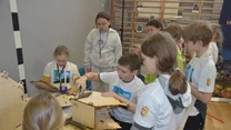 Grupa dzieci skupionych wokół stołu konstruuje model z drewnianych elementów, w tle nauczycielka nadzoruje zajęcia w sali gimnastycznej.