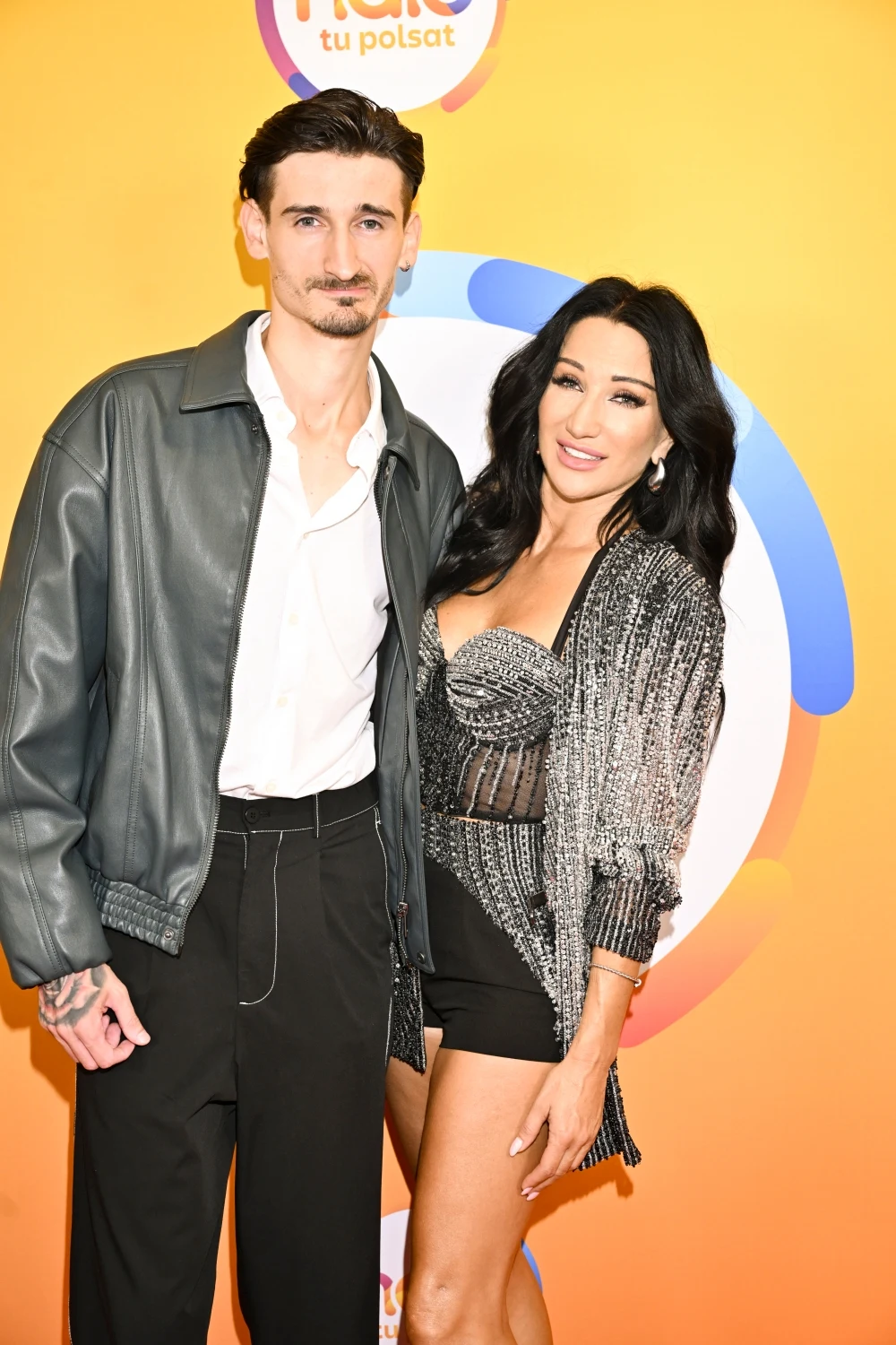 Dwie elegancko ubrane osoby pozują wspólnie na tle kolorowej ścianki z logiem Polsat. Mężczyzna w czarnej skórzanej kurtce i białej koszuli, kobieta w srebrnej, błyszczącej marynarce oraz krótkiej czarnej sukience.