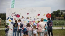 Grupa dzieci i dorosłych zgromadzona przed dużą tablicą, na której rysują kolorowe kwiaty, serca oraz napisy. W tle widoczna zieleń i fragmenty budynków. Słowo „lepsza” napisane dużymi literami.