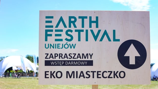 Tablica informacyjna z napisem Earth Festival Uniejów, zapraszająca do darmowego wstępu na eko miasteczko, w tle jasne namioty i trawiasta przestrzeń na świeżym powietrzu.