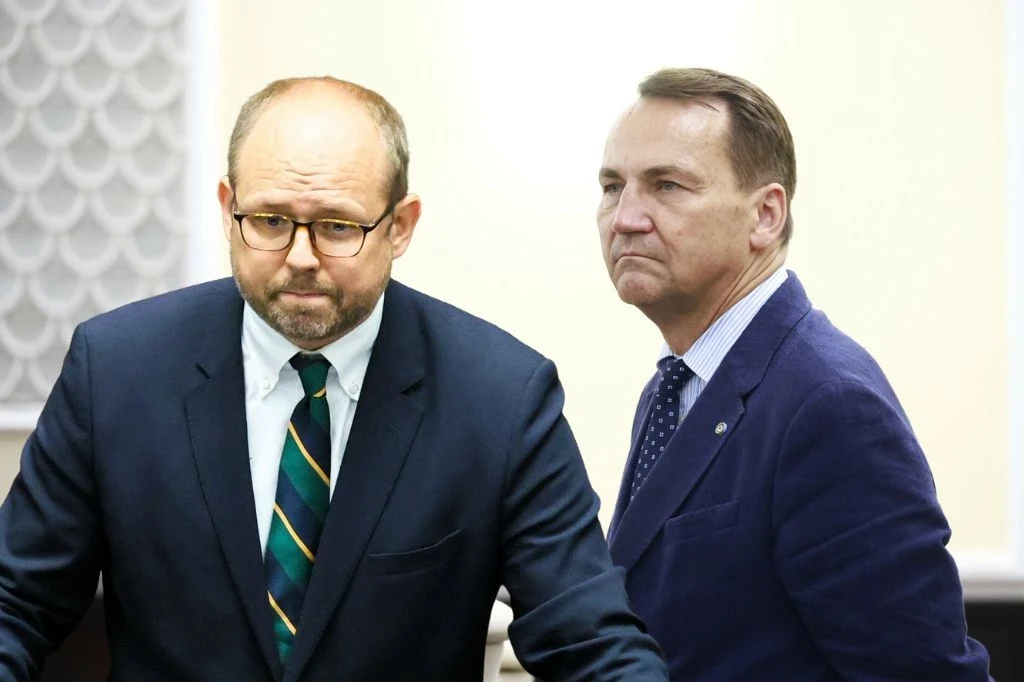 Zamieszanie wokół notatki ze spotkania w USA. Marcin Przydacz odpowiada Radosławowi Sikorskiemu Dwóch mężczyzn w garniturach stoi obok siebie w eleganckim, urzędowym pomieszczeniu. Jeden z nich ma okulary i pasiasty krawat, drugi ubrany w niebieską marynarkę, obaj mają poważne miny.