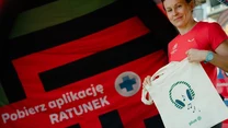Uśmiechnięta kobieta w czerwonej koszulce trzyma torbę z nadrukiem przy stoisku z dużym banerem zachęcającym do pobrania aplikacji Ratunek.