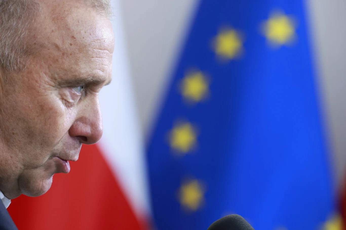 Mężczyzna w średnim wieku w profilu, przemawia do mikrofonu, w tle flaga Polski oraz flaga Unii Europejskiej w delikatnym rozmyciu.